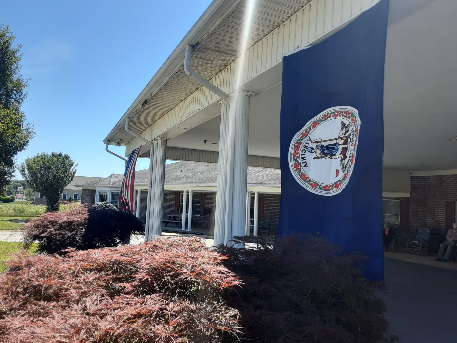 Exterior view of Birch Gardens Assisted Living showing a covered entrance with white columns, an American flag, and a Virginia state flag hanging. There are bushes and trees in the foreground under a clear blue sky.