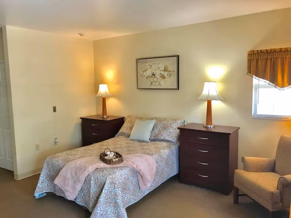 A cozy bedroom with a double bed covered in a patterned bedspread and a pink throw blanket. There are two dark wooden nightstands on either side of the bed, each with a lamp. A framed floral painting hangs above the bed. To the right, there is a window with a brown valance and a cushioned armchair next to it.