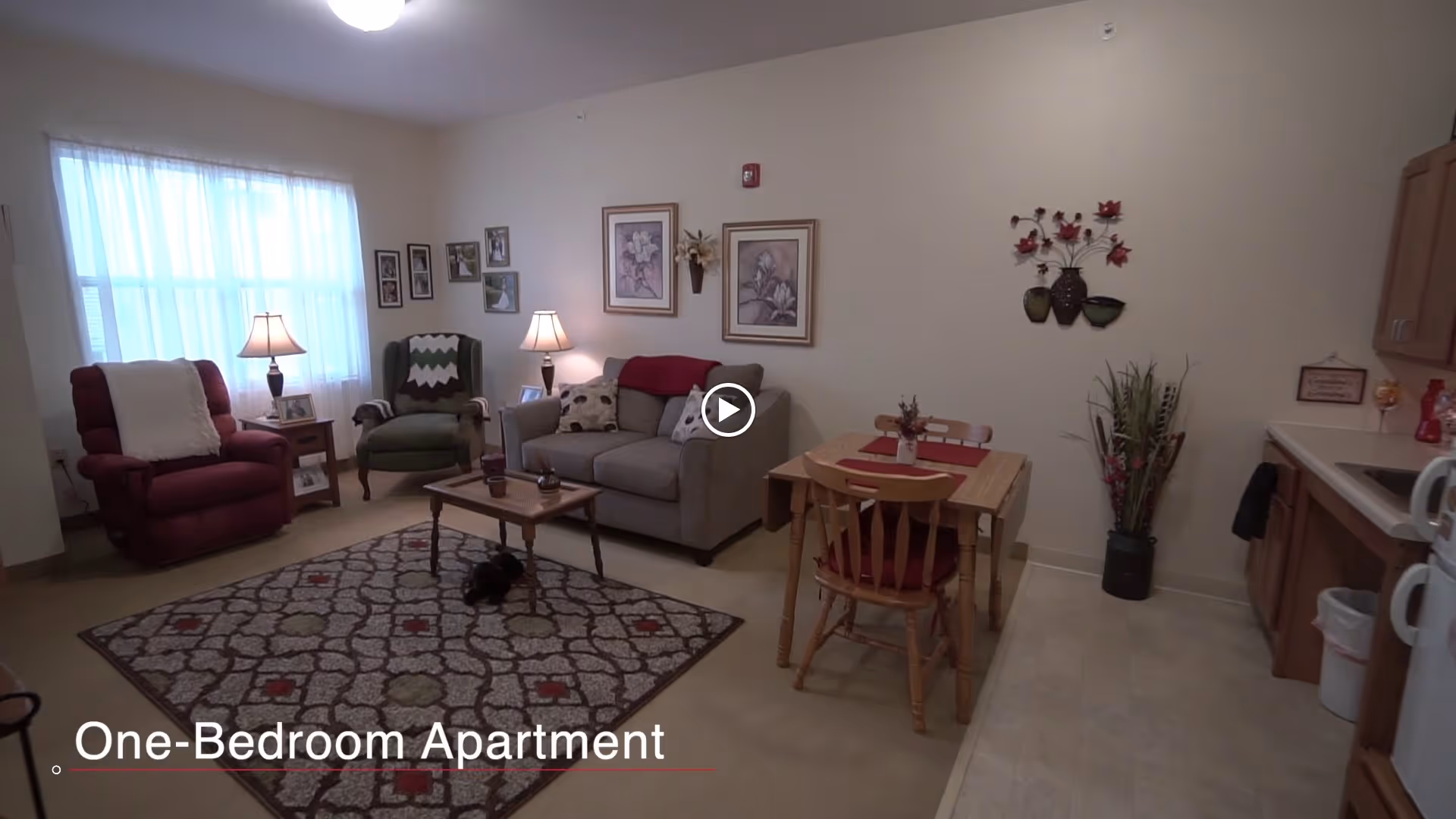 A cozy living area of a one-bedroom apartment featuring a red recliner, a green armchair, a gray sofa, a wooden coffee table on a patterned rug, a small wooden dining table with two chairs, and a kitchenette with wooden cabinets and a white microwave. The walls are decorated with framed pictures and floral wall art, and a window with sheer curtains allows natural light into the room.