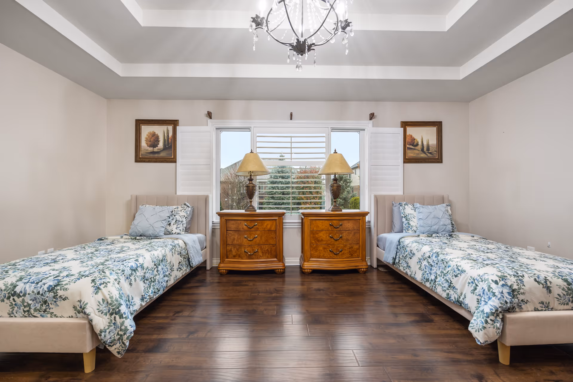 A bedroom with two twin beds on either side of a window with white shutters. Each bed has floral bedding in shades of blue and green, with matching pillows. Between the beds are two wooden nightstands with three drawers each, topped with matching table lamps. The room has wooden flooring, beige walls, and a decorative chandelier hanging from the ceiling. Two framed landscape paintings hang on the wall above each bed.