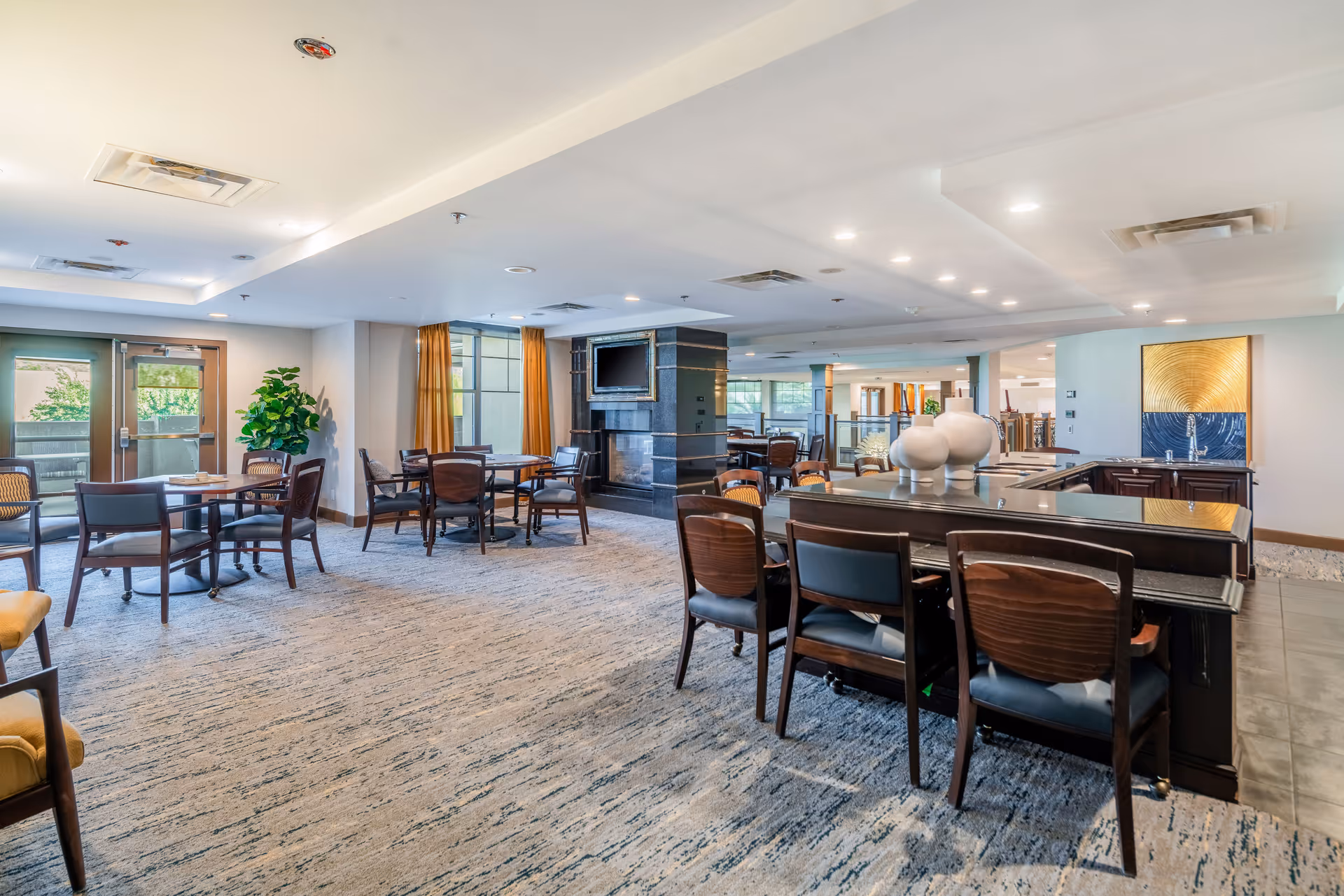 A spacious and well-lit common area with multiple tables and chairs arranged for seating. The room features a carpeted floor, large windows with orange curtains, a fireplace with a mounted TV above it, and a counter with decorative vases. The ceiling has recessed lighting and ventilation fixtures.