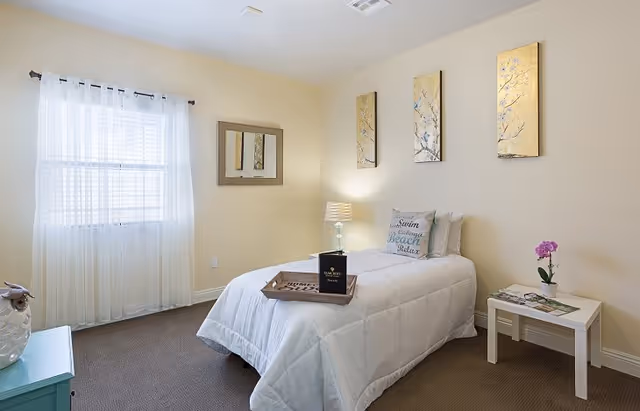 A cozy bedroom with a single bed covered in a white comforter and decorative pillows. A wooden tray with a book and a box is placed on the bed. The room has beige walls with three vertical floral paintings above the bed. A small white side table with a purple orchid and magazines is next to the bed. A window with sheer white curtains lets in natural light, and a mirror hangs on the wall beside the window. The floor is carpeted in a neutral tone.