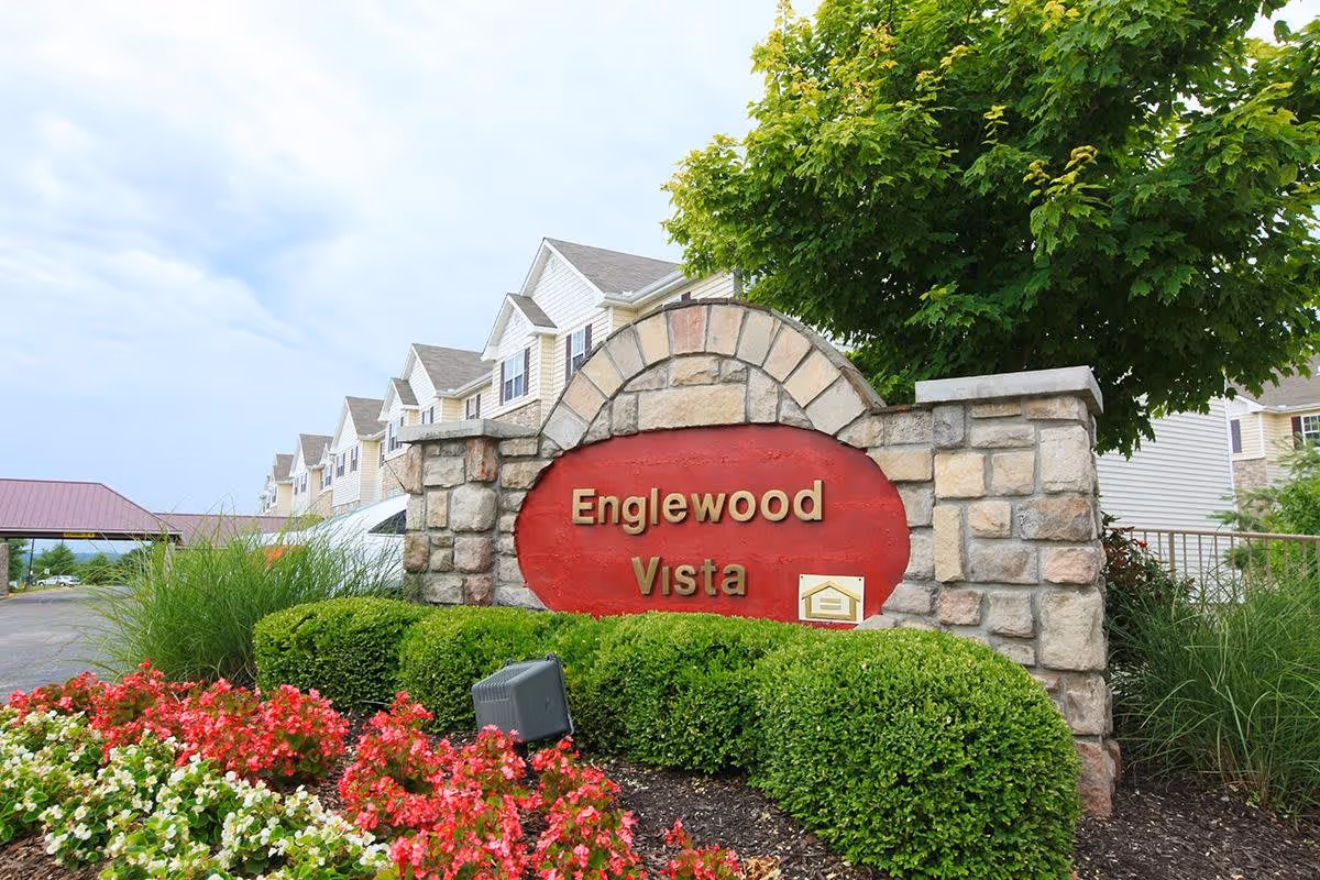 Stone and brick entrance sign for Englewood Vista surrounded by green bushes and colorful flowers, with a multi-story residential building and a tree in the background under a cloudy sky.
