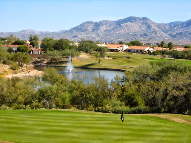 View of a golf course green and flag with a pond fountain, surrounding homes, desert vegetation, and mountains in the background.