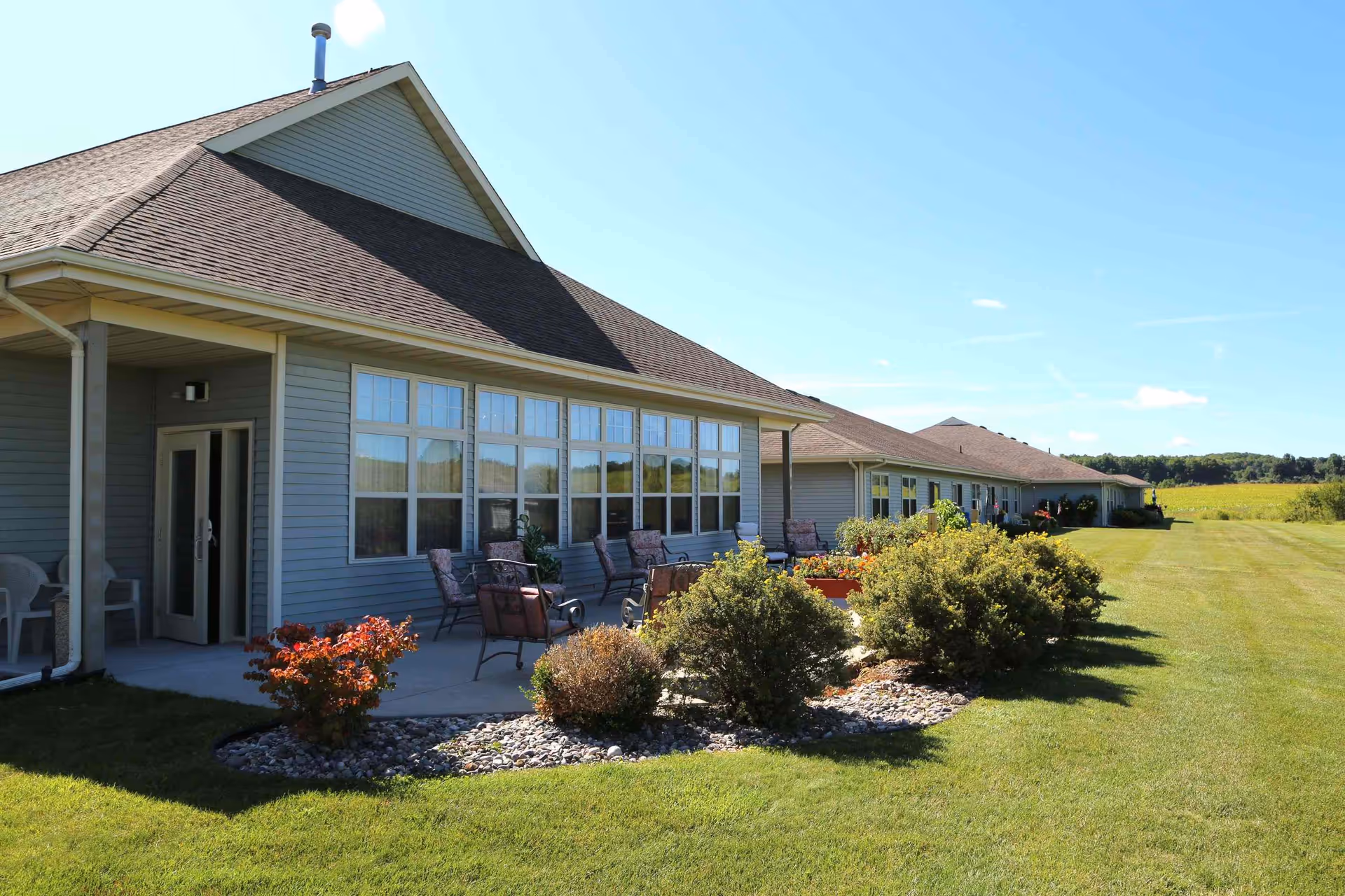 Exterior view of a single-story building with a sloped roof and multiple windows. There is a patio area with several chairs and small tables, surrounded by landscaped bushes and plants. The building is set against a backdrop of a large grassy field and clear blue sky.