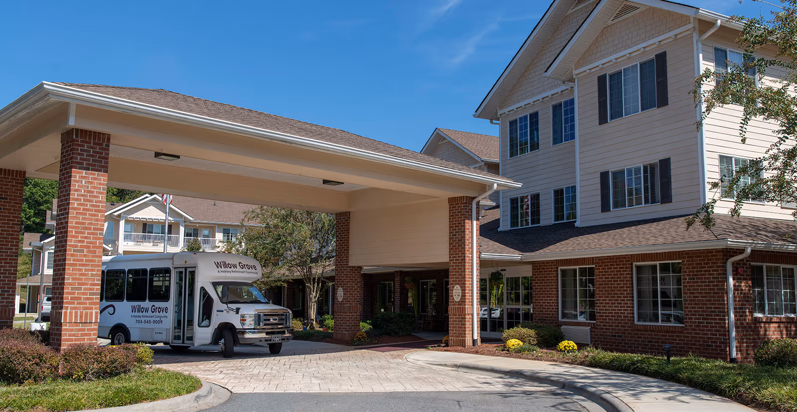Exterior view of Holiday Willow Grove senior living facility showing a covered entrance with brick pillars and a white shuttle bus parked underneath. The building has multiple windows and a well-maintained landscape with bushes and flowers under a clear blue sky.