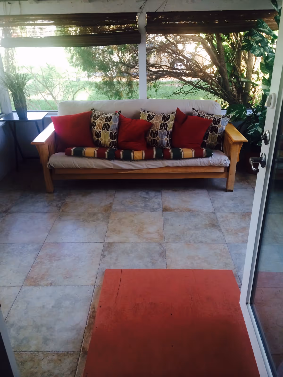 Wooden sofa with patterned and red throw pillows in a tiled sunroom/porch with plants visible outside the windows.