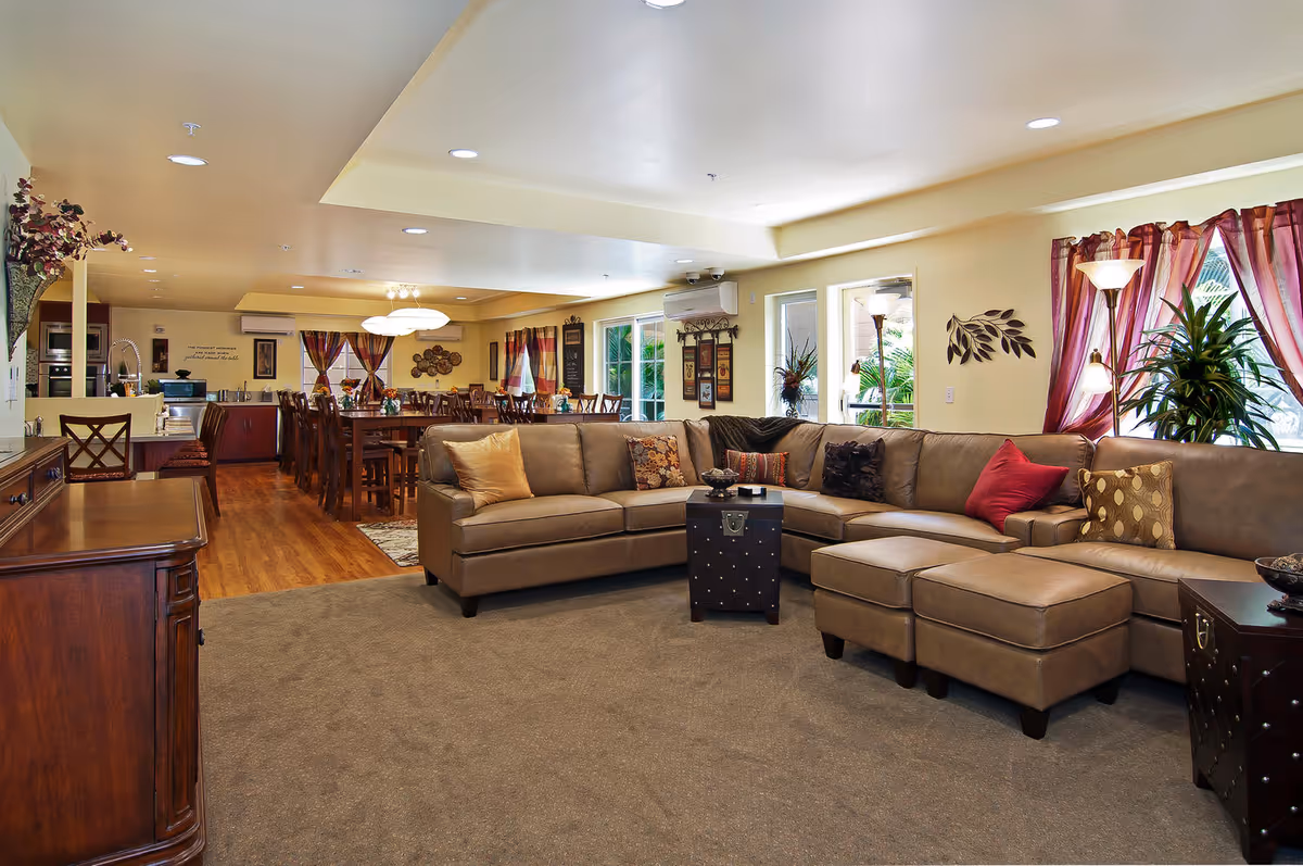 Spacious senior living common area with a large beige sectional sofa adorned with various decorative pillows, a small black coffee table, and two ottomans. The room features carpeted flooring in the seating area and wood flooring in the dining and kitchen areas. The dining area has a long wooden table with multiple chairs, and the kitchen is equipped with modern appliances. Large windows with sheer red curtains allow natural light to fill the space, complemented by ceiling lights and floor lamps. Decorative plants and wall art add warmth to the room.