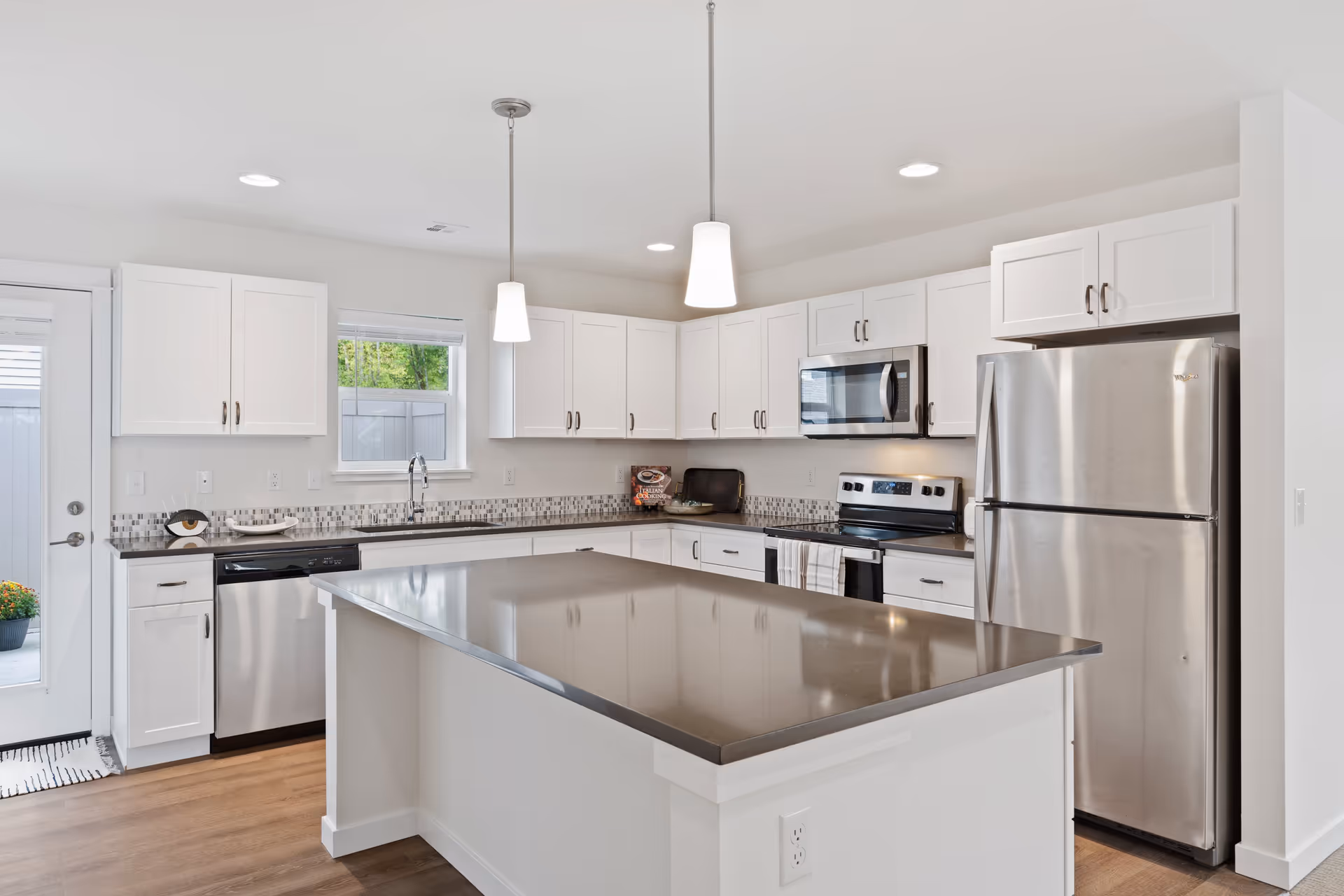 Modern kitchen with white cabinets, stainless steel appliances including a refrigerator, microwave, dishwasher, and electric stove. There is a large kitchen island with a smooth countertop in the center, two pendant lights hanging from the ceiling, and a small window above the sink letting in natural light.