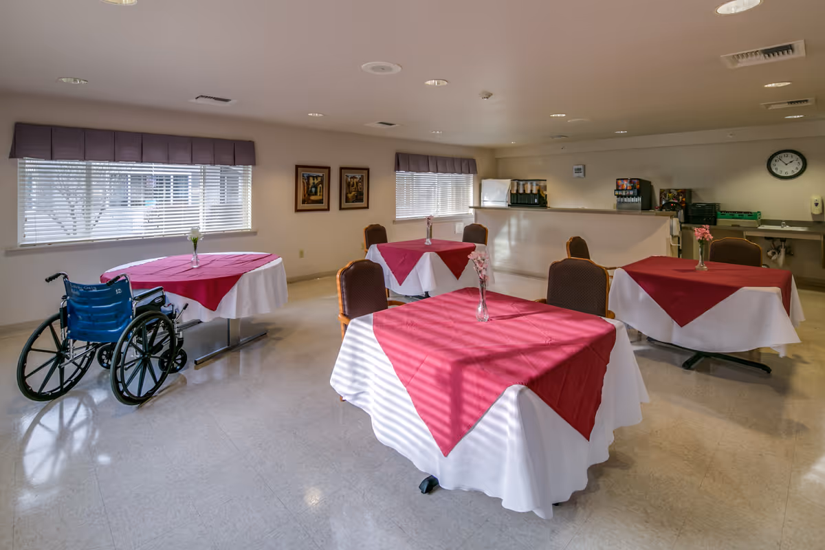 A dining room with four tables covered in white tablecloths and red table runners, each with a small vase of flowers. There are chairs around the tables and a blue wheelchair next to one table. The room has two windows with blinds and purple valances, a counter with beverage dispensers, and a clock on the wall.