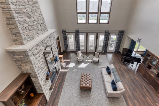 Spacious living room with high ceilings, large stone fireplace, beige sofa with blue pillows, two armchairs with a small round table between them, a tufted ottoman, dark wood flooring, large windows with blue patterned curtains, and a black grand piano near a wooden cabinet.