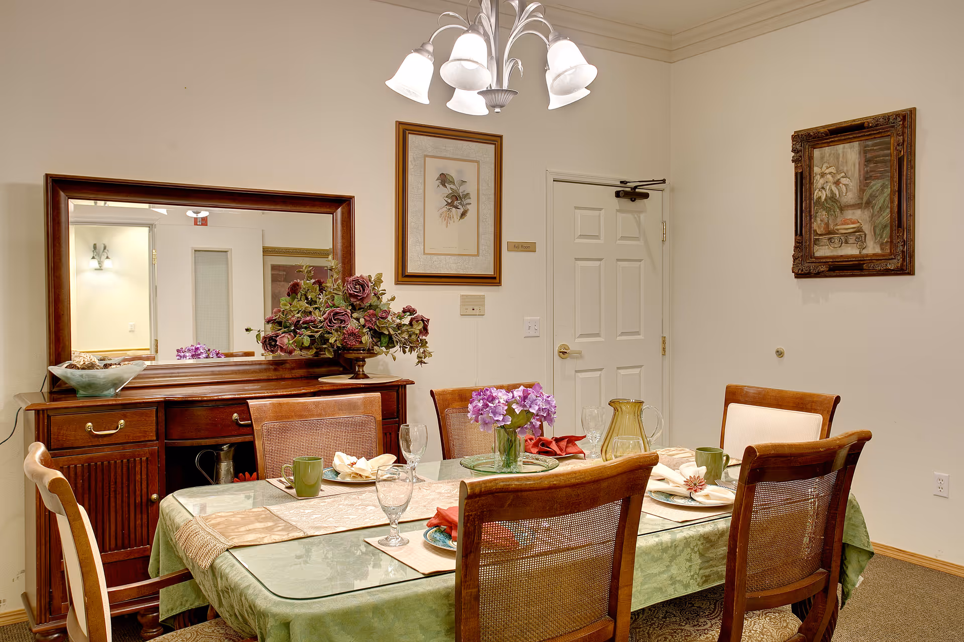 A dining room with a rectangular glass-top table covered with a green tablecloth and set with plates, glasses, and napkins. Six wooden chairs with woven backs surround the table. A wooden sideboard with a large mirror and a floral arrangement sits against the wall. Two framed pictures hang on the walls, and a chandelier with multiple lights hangs above the table.