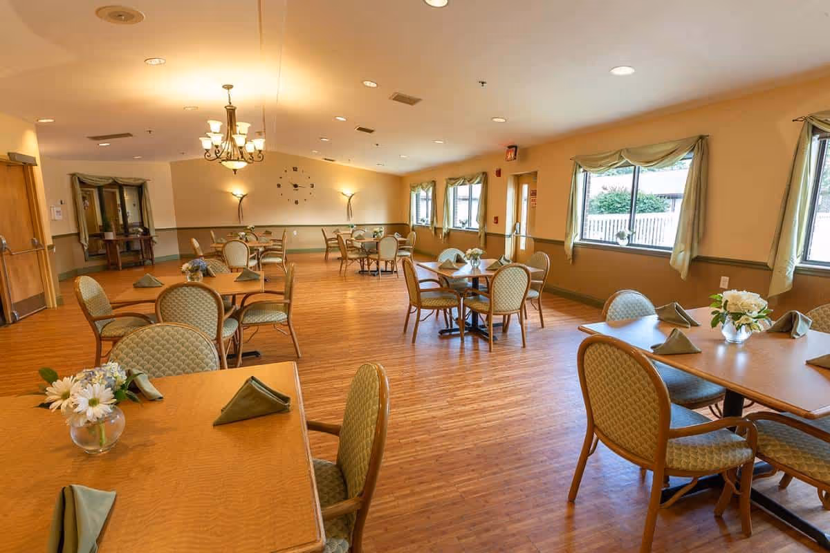 Bright senior living dining room with several round and rectangular tables, cushioned chairs, wooden floors and windows.