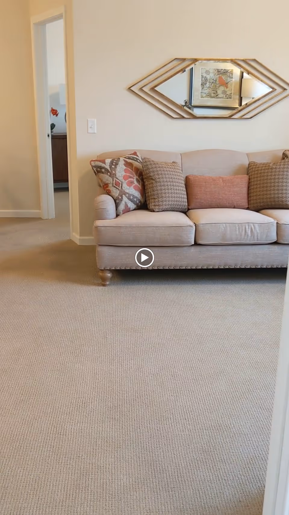 A beige sofa with four decorative pillows in various patterns and colors is placed against a light-colored wall. Above the sofa hangs a diamond-shaped mirror with a wooden frame. To the left, there is an open doorway leading to another room with a cabinet and a red flower visible. The floor is carpeted in a light beige color.
