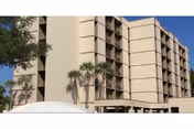 Beige multi-story apartment building with balconies, palm trees, and a covered entrance under a clear blue sky.