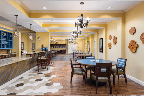 A spacious dining area with wooden floors and hexagonal tile accents. There are round and rectangular tables with chairs arranged neatly. The walls are painted yellow and decorated with wooden wall art. Multiple chandeliers and pendant lights hang from the ceiling, providing warm lighting. A long counter with bar stools is visible on the left side.