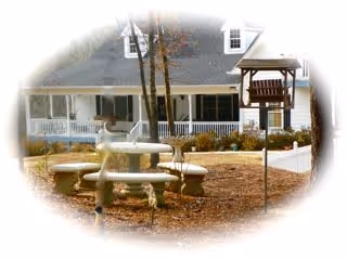 Outdoor area of a senior living facility named Olive Branch featuring a round stone picnic table with attached benches, a bird feeder on a pole, and a large white building with a porch and multiple windows in the background. Trees and dry leaves are visible on the ground.