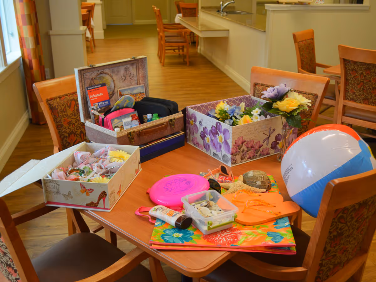 A wooden table in a communal dining or activity area with various items on it including a beach ball, orange flip-flops, a pink frisbee, a box of seashells, a tube of sunscreen, and decorative boxes filled with artificial flowers and travel-related items. The room has wooden chairs with patterned cushions and a kitchen area visible in the background.