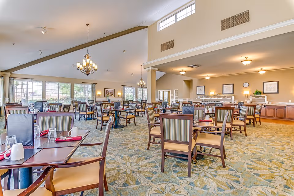 Spacious dining room in a senior living facility with multiple wooden tables and chairs arranged neatly. The room features large windows letting in natural light, chandeliers hanging from a high ceiling, and a carpeted floor with a floral pattern. The tables are set with glasses, mugs, and red napkins.