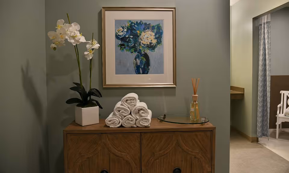 A wooden cabinet with a white orchid plant in a square pot, a stack of rolled white towels arranged in a pyramid, and a glass tray with a reed diffuser on top. Above the cabinet hangs a framed painting of a blue vase with white and blue flowers. To the right, there is a partial view of a room with a white chair and a curtain with a chevron pattern.