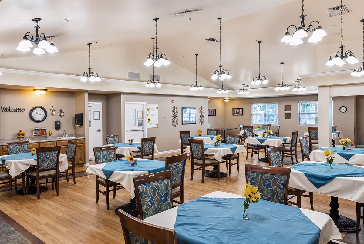 A spacious dining room with multiple tables covered in white tablecloths and blue table runners, each decorated with a small vase of yellow flowers. The room has wooden chairs with patterned upholstery, hardwood flooring, and multiple hanging light fixtures. There are windows letting in natural light and a wall clock with a 'Welcome' sign on one wall.