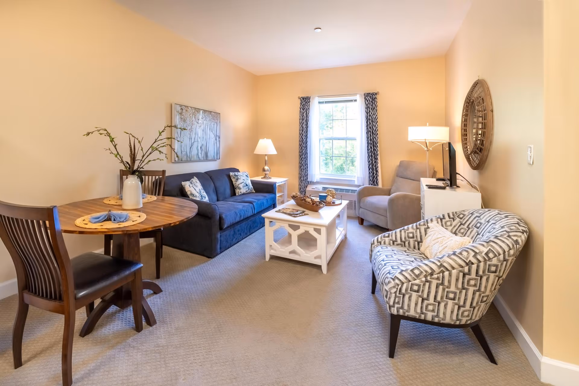 Well-lit living room with a sofa, patterned armchair, round dining table, coffee table, and a window.