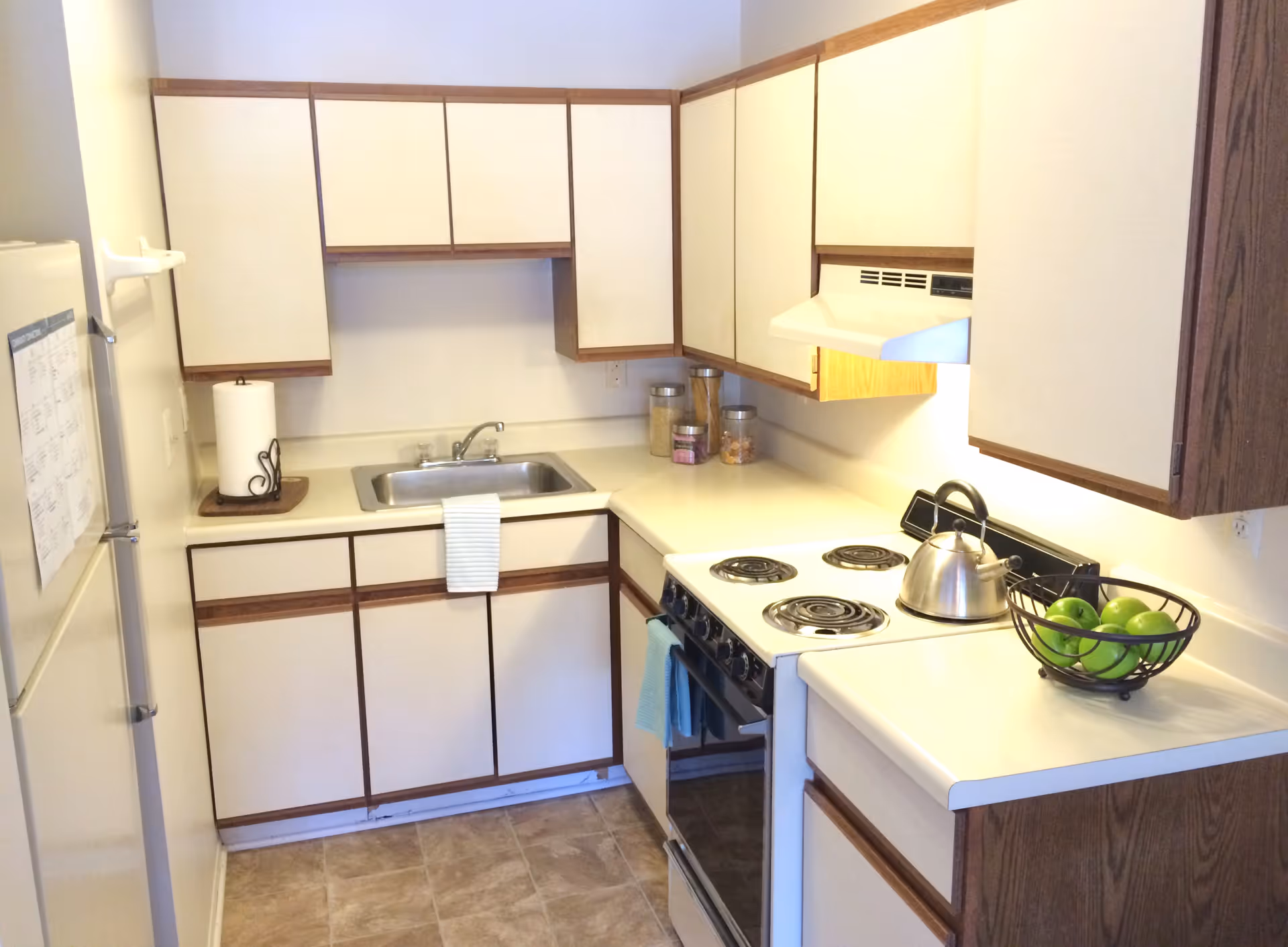 A compact kitchen with cream-colored cabinets and wood trim, a stainless steel sink with a striped towel hanging on the cabinet handle, a white electric stove with a kettle on one burner, a bowl of green apples on the counter, a paper towel holder, and three jars with food items on the countertop.