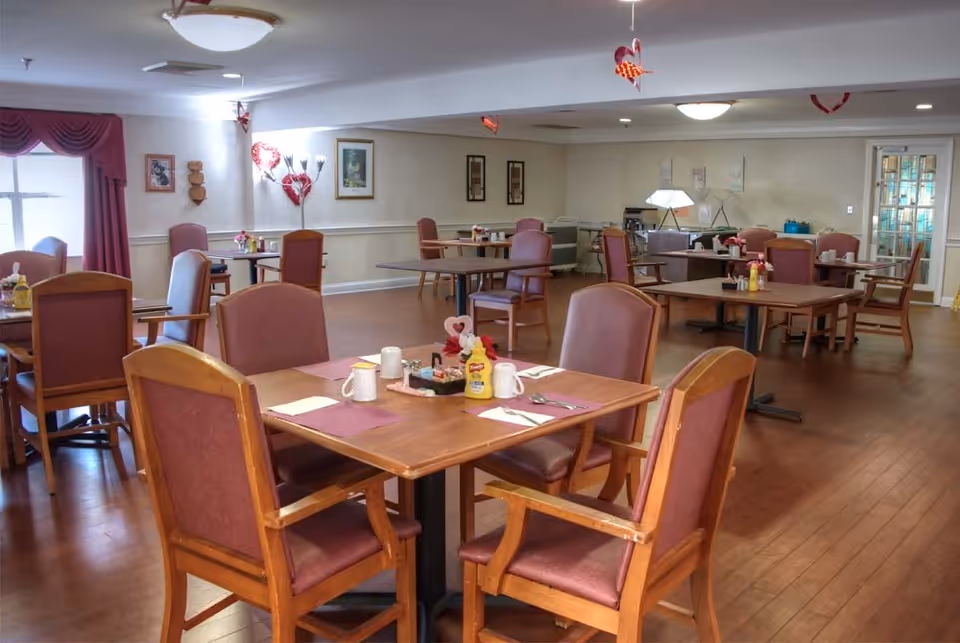 A dining room in an assisted living facility with several wooden tables and chairs arranged neatly. Each table is set with placemats, napkins, mugs, and condiments. The room has hardwood floors, light-colored walls with framed pictures, and windows with red curtains. Ceiling lights illuminate the space, and there are some small decorative items hanging from the ceiling.