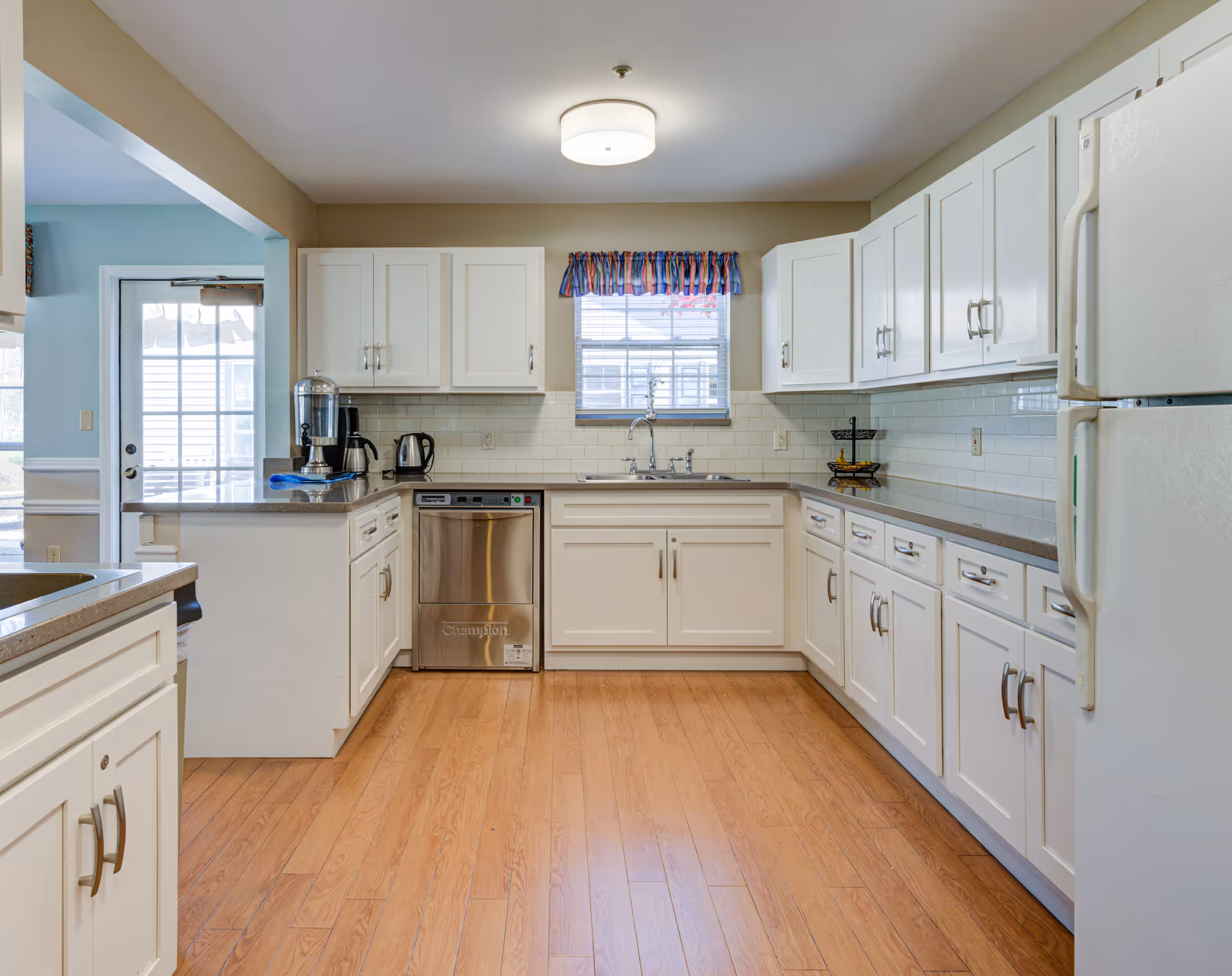 A clean, well-lit kitchen with white cabinets, a stainless steel dishwasher, a white refrigerator, and a window with a colorful valance above the sink. The kitchen has light wood flooring and beige walls, with a countertop holding a water dispenser, coffee maker, and electric kettle.