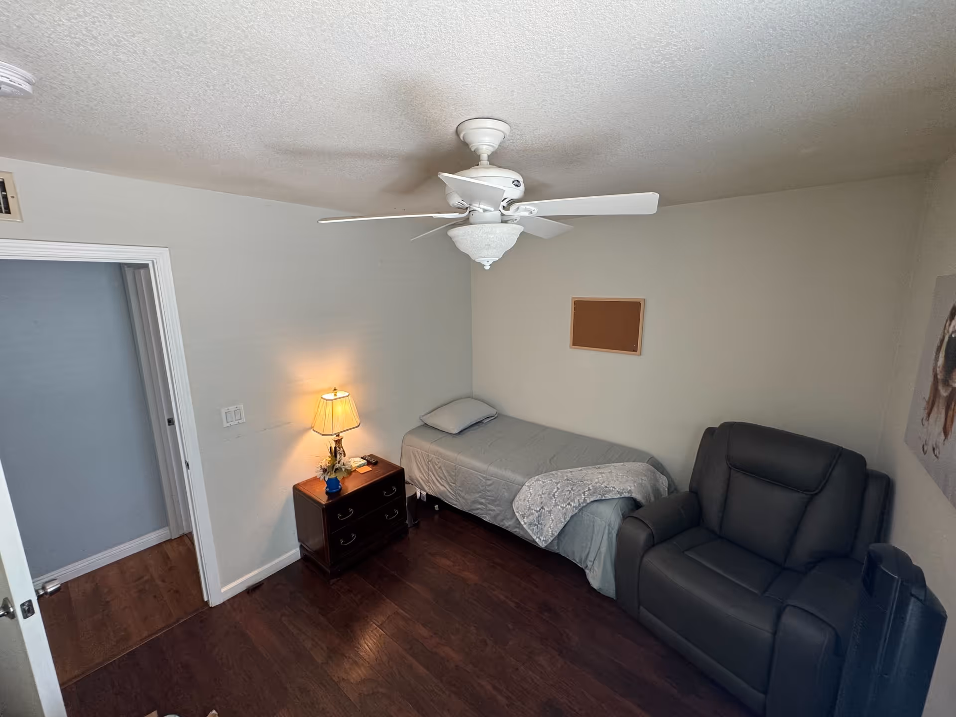 A small bedroom with a single bed covered with gray bedding, a wooden nightstand with a lamp and small decorative items, a dark gray recliner chair, a ceiling fan with light, and a corkboard on the wall. The room has wooden flooring and light-colored walls.