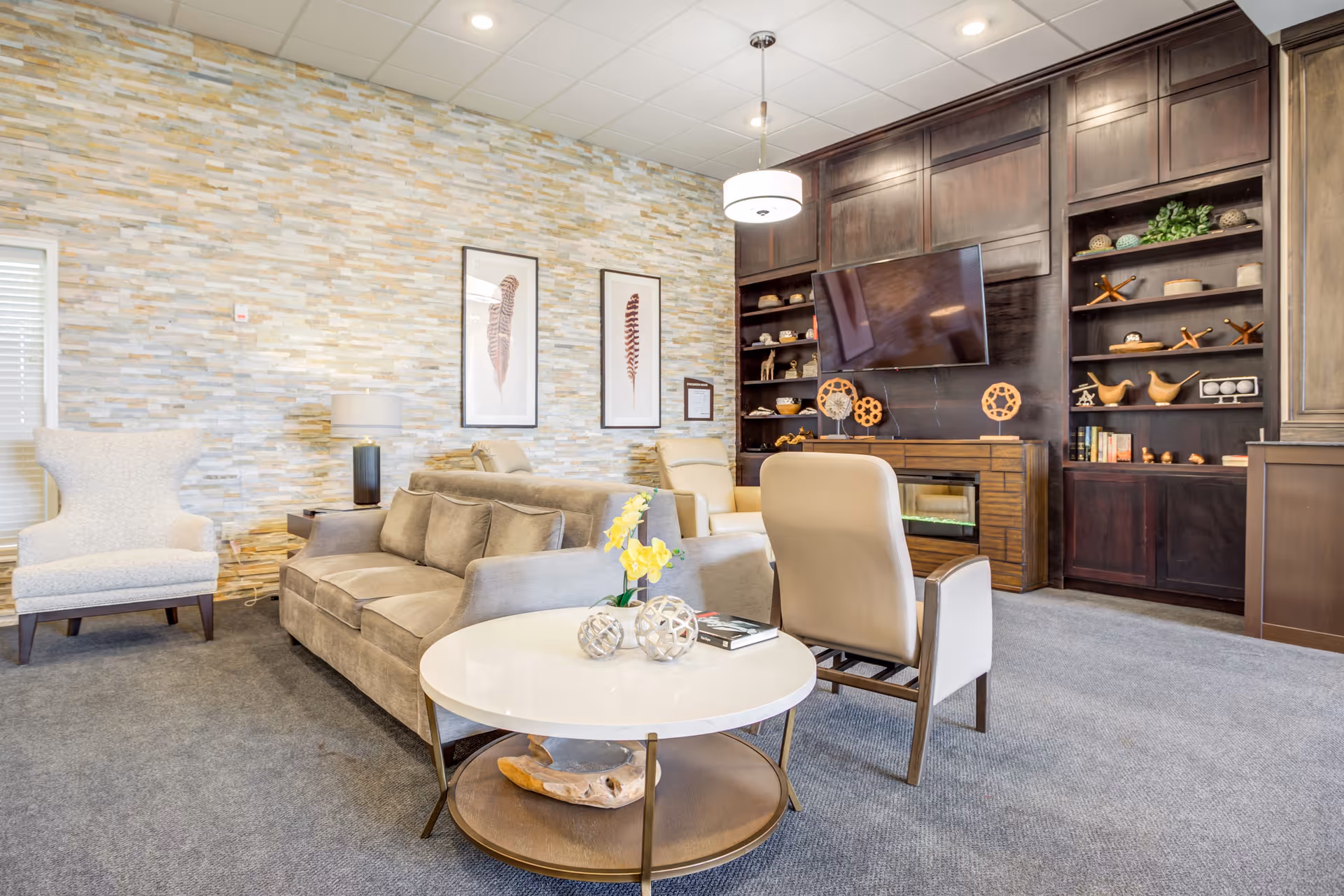 Modern communal living room with sofas, armchairs, a round coffee table, and built-in dark wood shelving with a mounted TV.