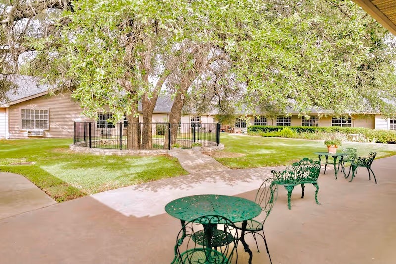 Outdoor patio area with green metal tables and chairs under a covered walkway. In the background, there is a grassy courtyard with large trees surrounded by a black metal fence and beige buildings with multiple windows.