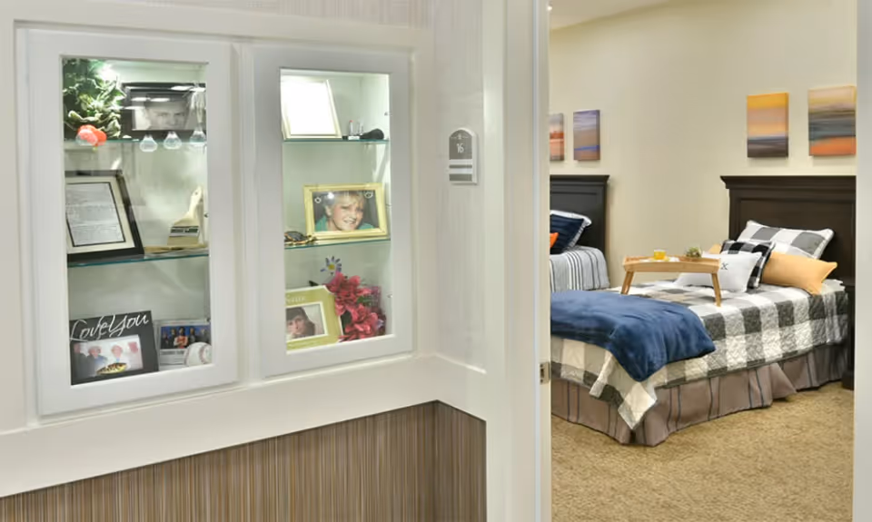 View into a senior living bedroom with two twin beds, each with checkered bedding and pillows. A wooden tray with breakfast items is on one bed. On the left, there is a built-in glass display cabinet with framed photos and decorative items. The room number 16 is visible on the wall near the door.