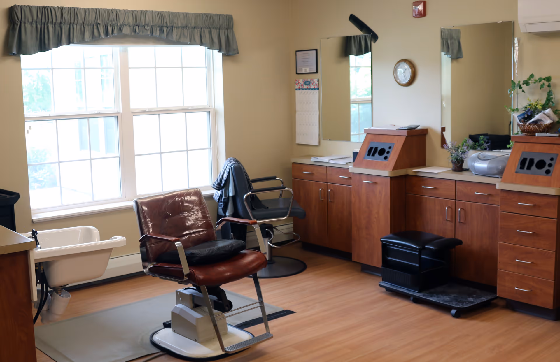 Salon-style grooming room with styling chairs, a wash basin, mirrors, and wooden cabinetry beneath a large window.
