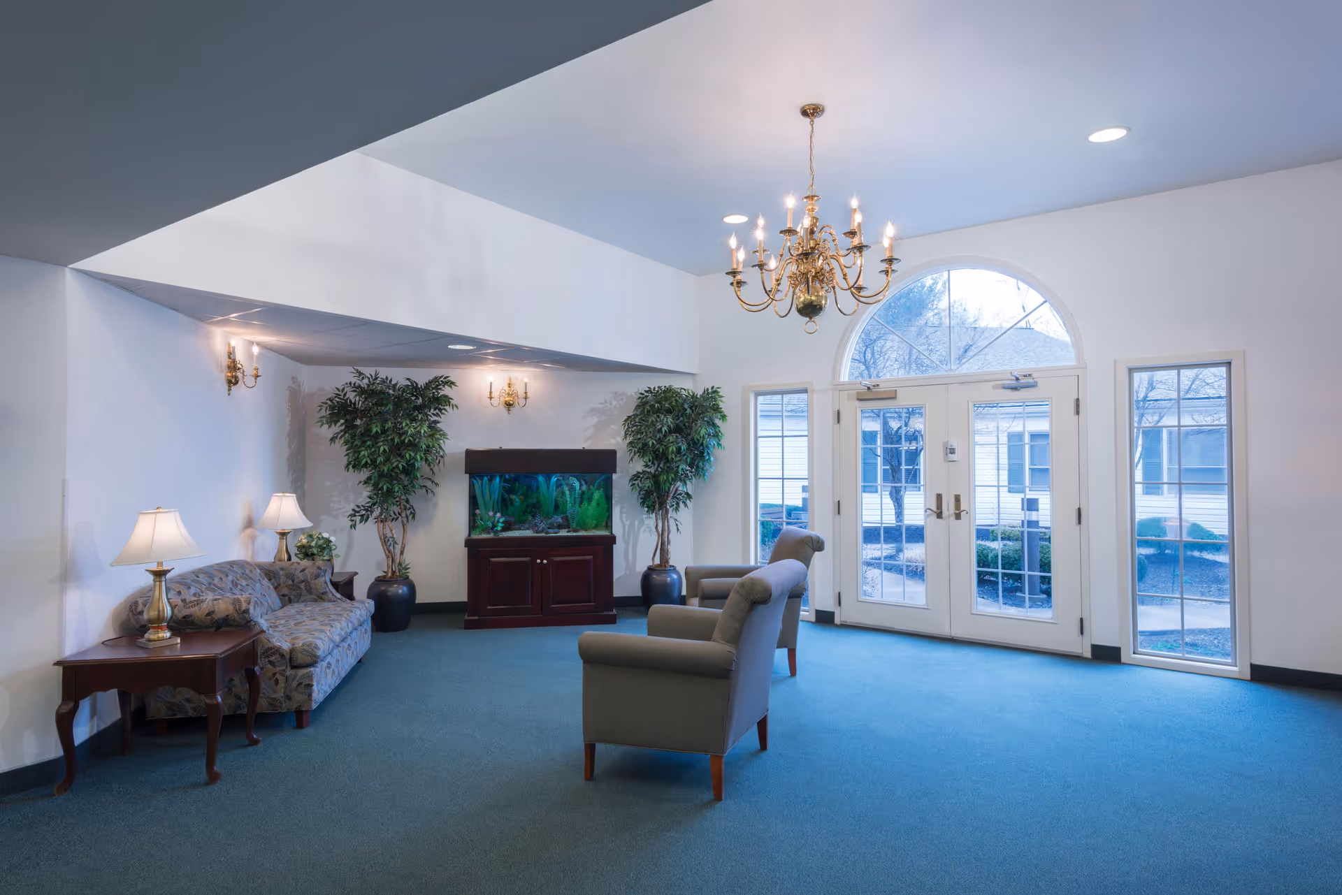 A bright and spacious common area with blue carpet, two armchairs facing a large windowed double door, a floral patterned sofa with side tables and lamps, two large potted plants, and a large fish tank on a wooden stand. A chandelier hangs from the ceiling, and wall sconces provide additional lighting.
