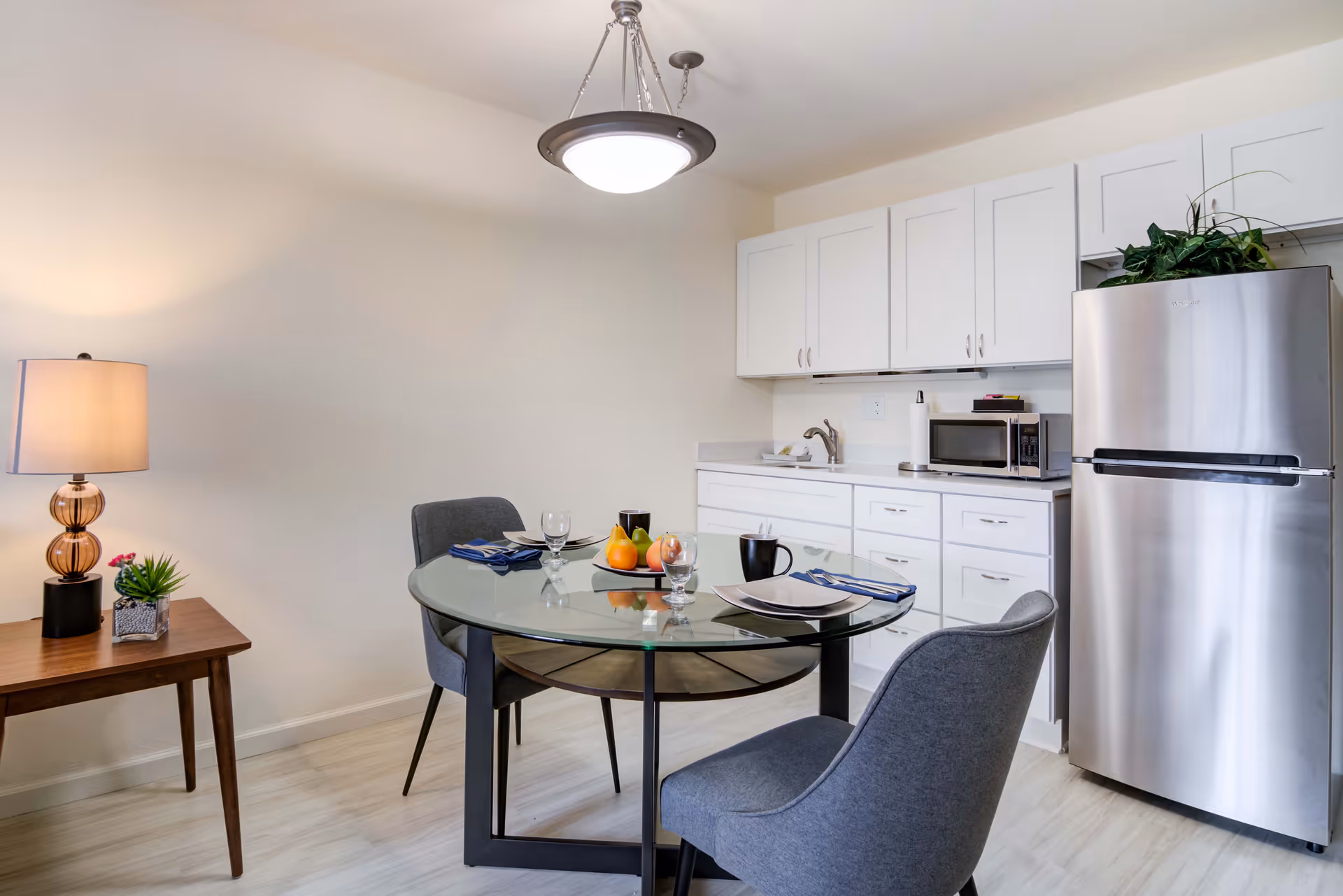Small dining area with a glass round table set for two, gray chairs, a lamp on a side table, and a kitchenette with white cabinets and a stainless steel refrigerator.