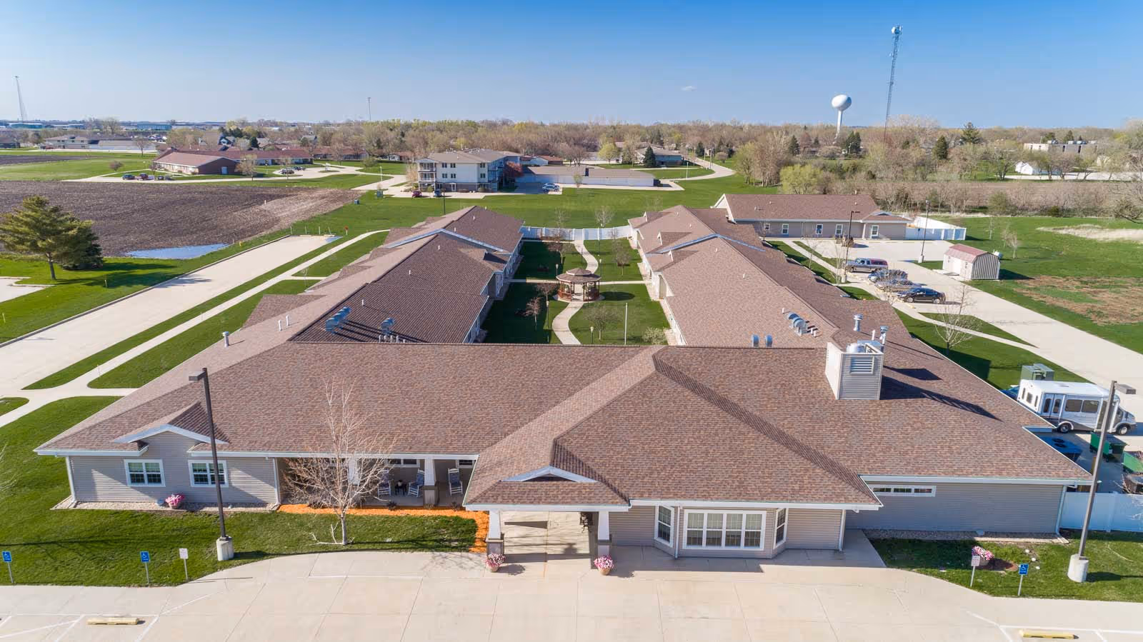 Aerial view of Windsor Manor senior living facility showing multiple connected buildings with brown roofs, green lawns, paved walkways, and a parking area in front. The surrounding area includes open fields and trees under a clear blue sky.