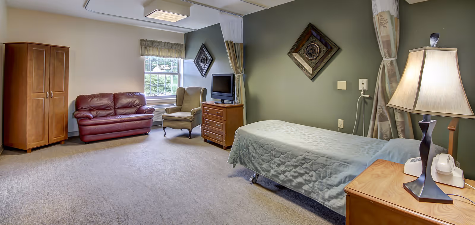 A senior living facility room with a single bed covered in a light blue quilt, a wooden nightstand with a lamp and telephone, a wooden dresser with a small TV on top, a beige armchair, a red leather loveseat, a wooden wardrobe, and a window with a valance letting in natural light. The walls are painted in two tones, beige and green, with decorative wall art.