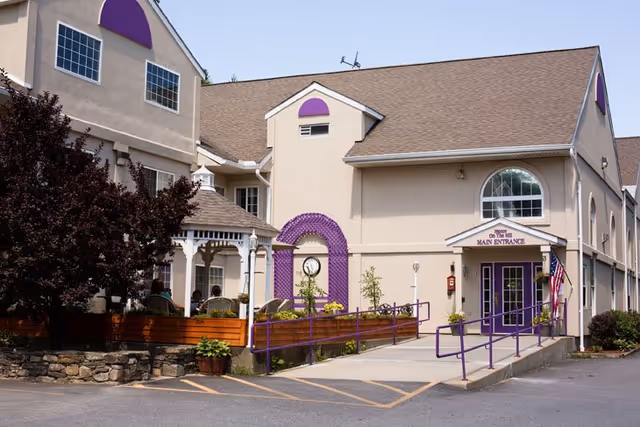 Exterior view of Manor On the Hill senior living facility showing the main entrance with a purple awning, a wheelchair accessible ramp with purple railings, a gazebo, and landscaping including trees and plants.