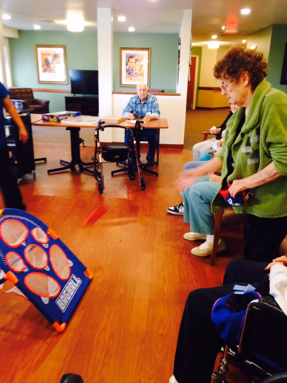 A group of elderly people in a common area of a senior living facility. One woman in a green sweater is playing a game of bean bag toss, aiming at a blue and orange target labeled 'BAGBALL'. Other residents are seated nearby, and a man sits at a table with a walker in front of him. The room has wooden floors, green walls, and framed pictures on the wall.