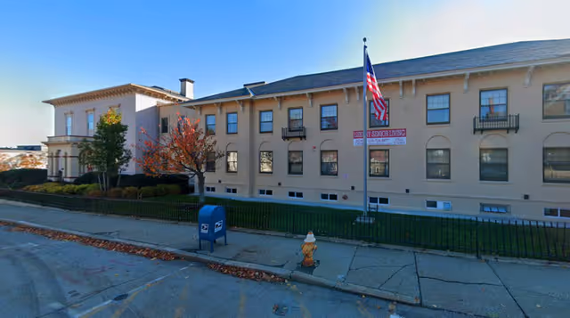 Exterior view of a two-story beige building with multiple windows and small balconies. There is a flagpole with an American flag in front of the building, along with a blue USPS mailbox and a yellow fire hydrant on the sidewalk. Trees with autumn foliage and a black metal fence line the front of the property.