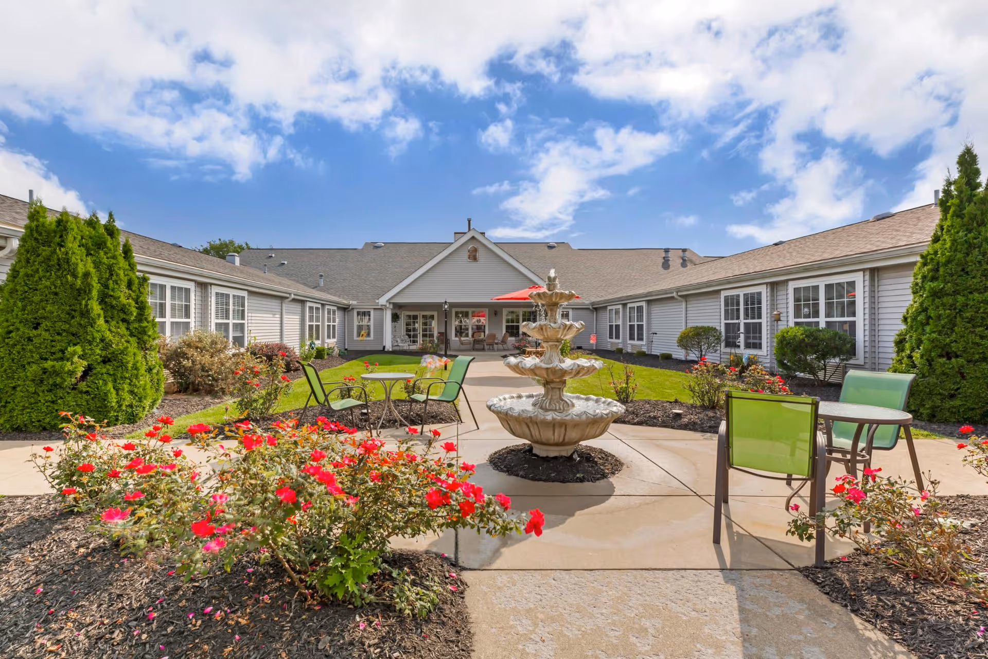 Sunny courtyard with a three-tier fountain, patio tables and chairs, red flowers, and a surrounding single-story building.