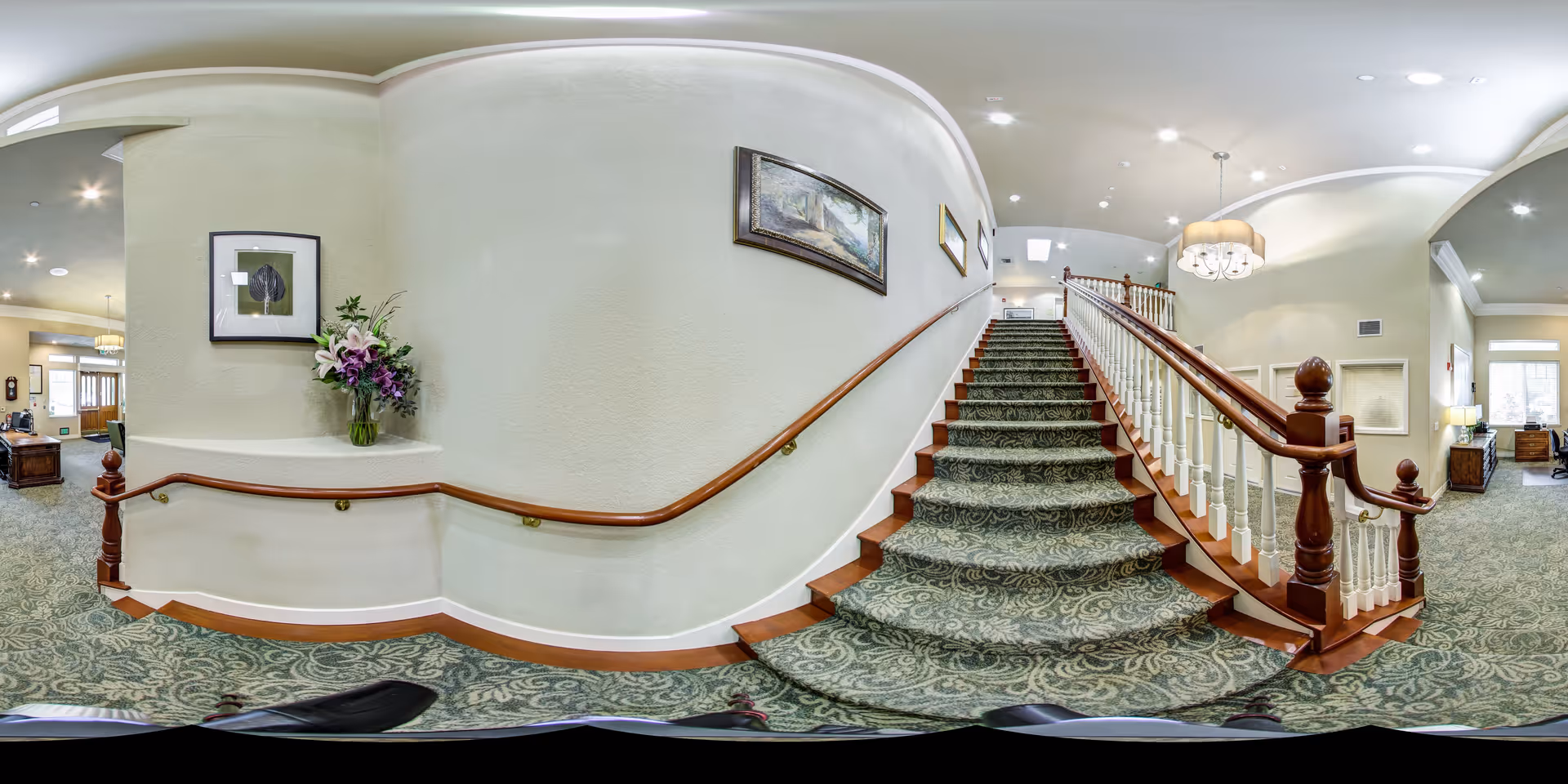 Interior view of a senior living facility showing a carpeted staircase with wooden handrails and white balusters. The walls are light-colored with framed artwork and a vase of flowers on a ledge. The area is well-lit with ceiling lights and a chandelier above the stairs. Adjacent rooms with furniture and windows are visible on either side of the staircase.