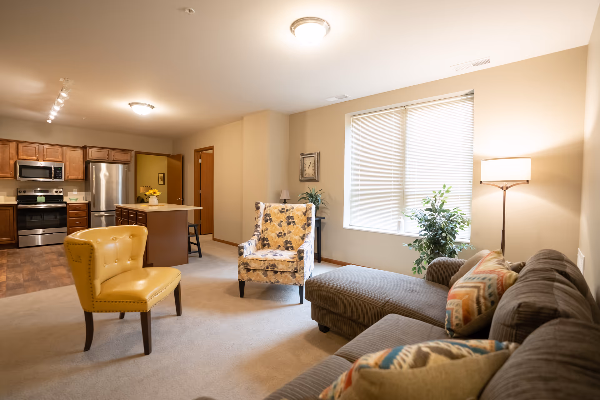 A bright and cozy living area featuring a gray sectional sofa with colorful patterned pillows, a floral armchair, and a yellow accent chair. The space includes a large window with blinds, a floor lamp, and a potted plant. In the background, there is a kitchen with wooden cabinets, stainless steel appliances, and an island with a stool.