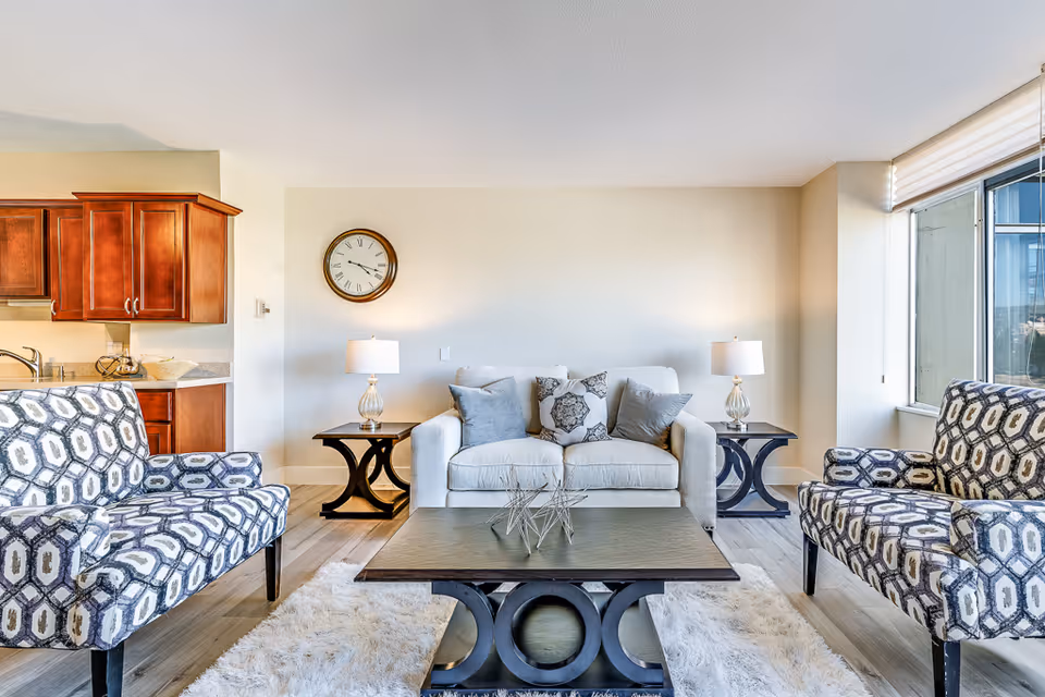 A bright and modern living room in a senior living facility with a white sofa adorned with decorative pillows, two patterned armchairs, a dark wooden coffee table, two matching side tables with lamps, a wall clock, and a view of a kitchen area with wooden cabinets on the left.