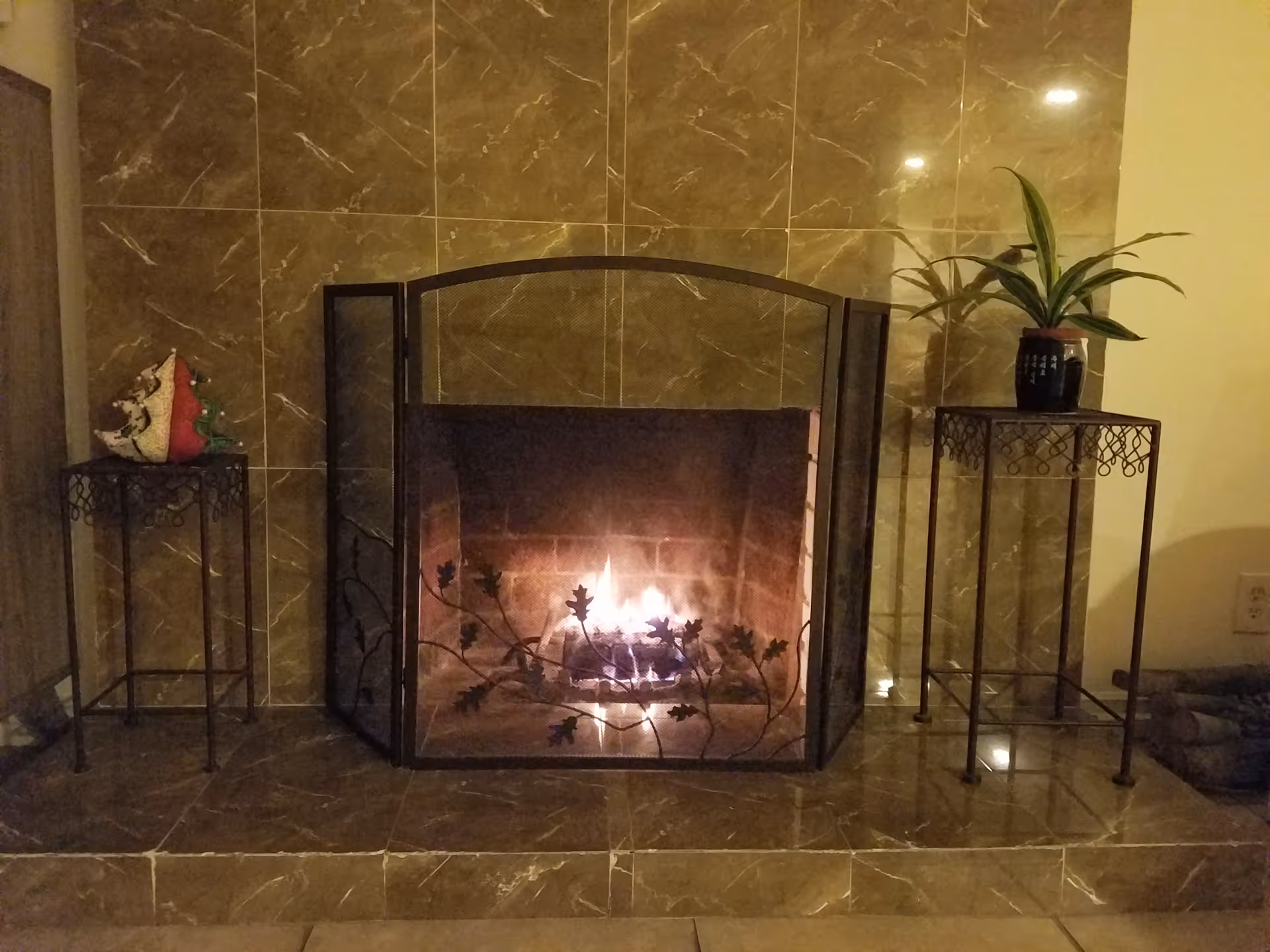 A lit fireplace with a decorative metal screen featuring leaf patterns. The fireplace is surrounded by brown marble tiles. On either side of the fireplace are small metal tables; the left table holds decorative items including a small Christmas tree, and the right table holds a potted plant. The setting appears cozy and warm.