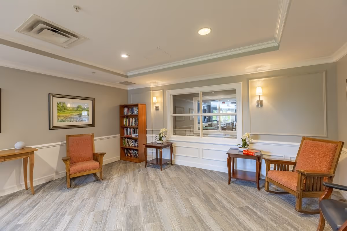 A cozy sitting area in a senior living facility with two wooden armchairs upholstered in orange fabric, two small wooden side tables with potted plants and books, a wooden bookshelf filled with books, a framed landscape painting on the wall, and a large window looking into another room. The room has light-colored walls, recessed ceiling lights, and wood-look flooring.