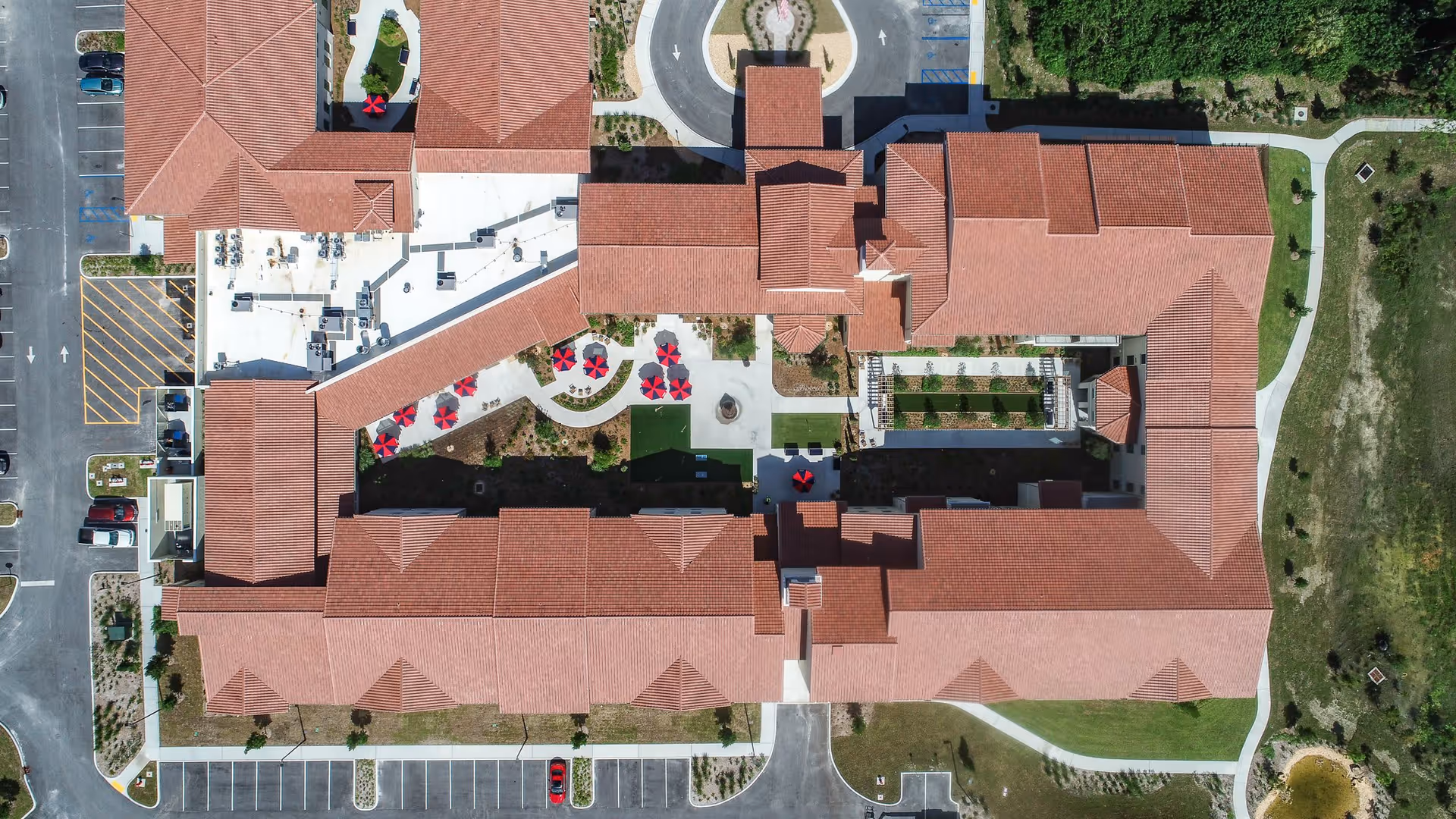 Aerial view of a U-shaped red-tiled roof senior living complex surrounding a landscaped central courtyard with walking paths, seating with red umbrellas, and adjacent parking.