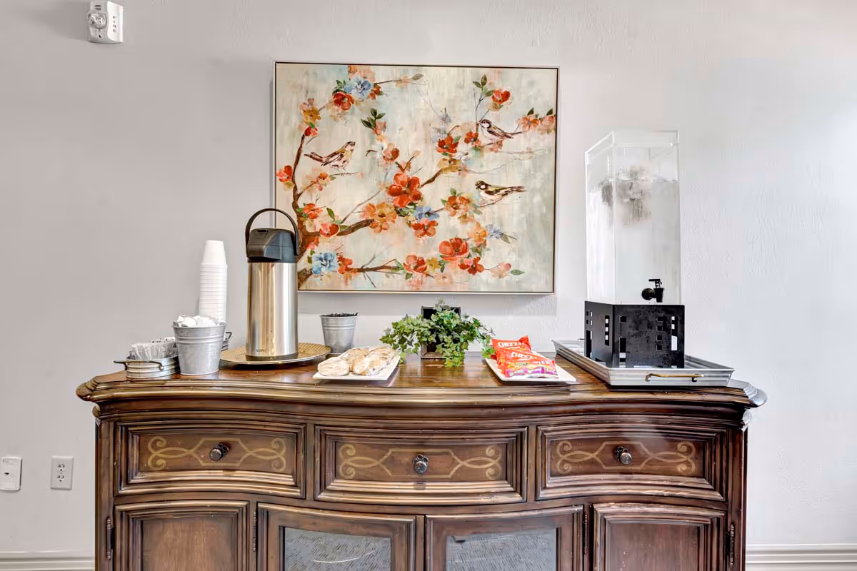 A wooden sideboard with three drawers and cabinets below, topped with a coffee dispenser, a stack of white disposable cups, a small bucket with stirrers, a tray of pastries, a small green potted plant, a tray with snack bags, and a large clear water dispenser with ice. Above the sideboard hangs a framed painting of birds perched on flowering branches with red, orange, and blue flowers.