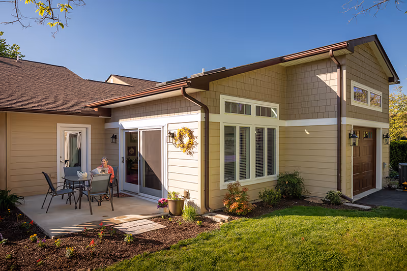 A sunny outdoor patio area of a residential facility with a woman sitting at a table with four chairs. The building has beige siding, white trim, a brown roof, and a garage door. There are plants and flowers around the patio and a green lawn in the foreground.